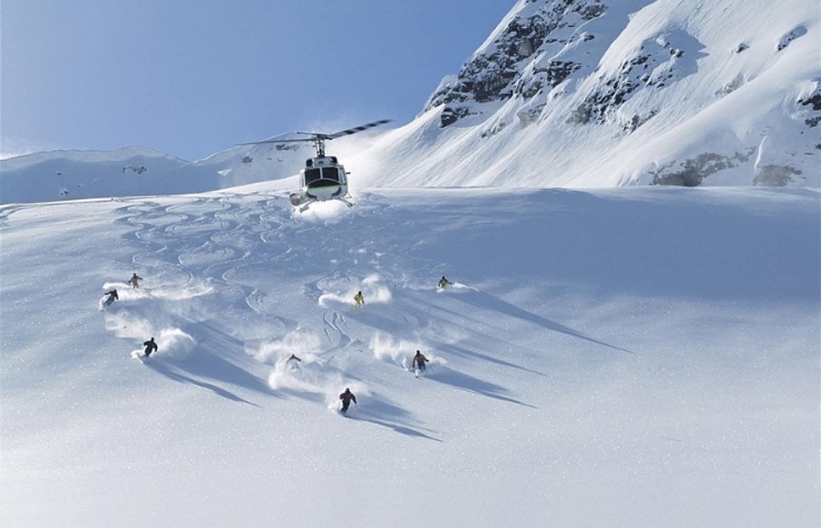 A panorama of skiers skiing through fresh powder snow with a helicopter flying above them